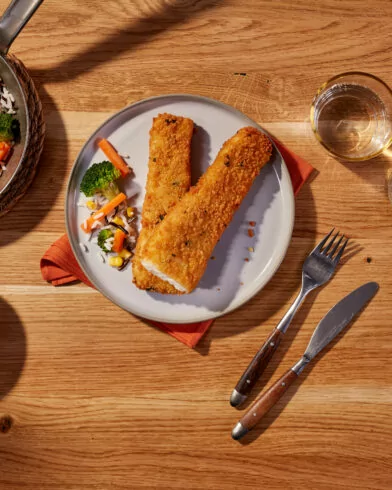 Egy tál, amelyen két FRoSTA rántott halfilé, FRoSTA vegyes zöldség, burgonyapüré, sárgarépás-kukoricás rizs, valamint két pohár fehérbor található.