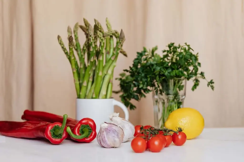 Friss zöldségek és fűszernövények, köztük spárga, petrezselyem, piros paprika, fokhagyma, koktélparadicsom és citrom fehér háttér előtt.