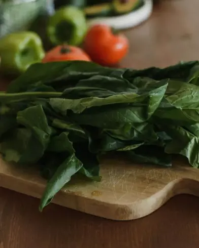Friss spenótlevelek, egy fa vágódeszkán, a háttérben elmosódott paradicsomokkal, zöldpaprikákkal és dióval.
